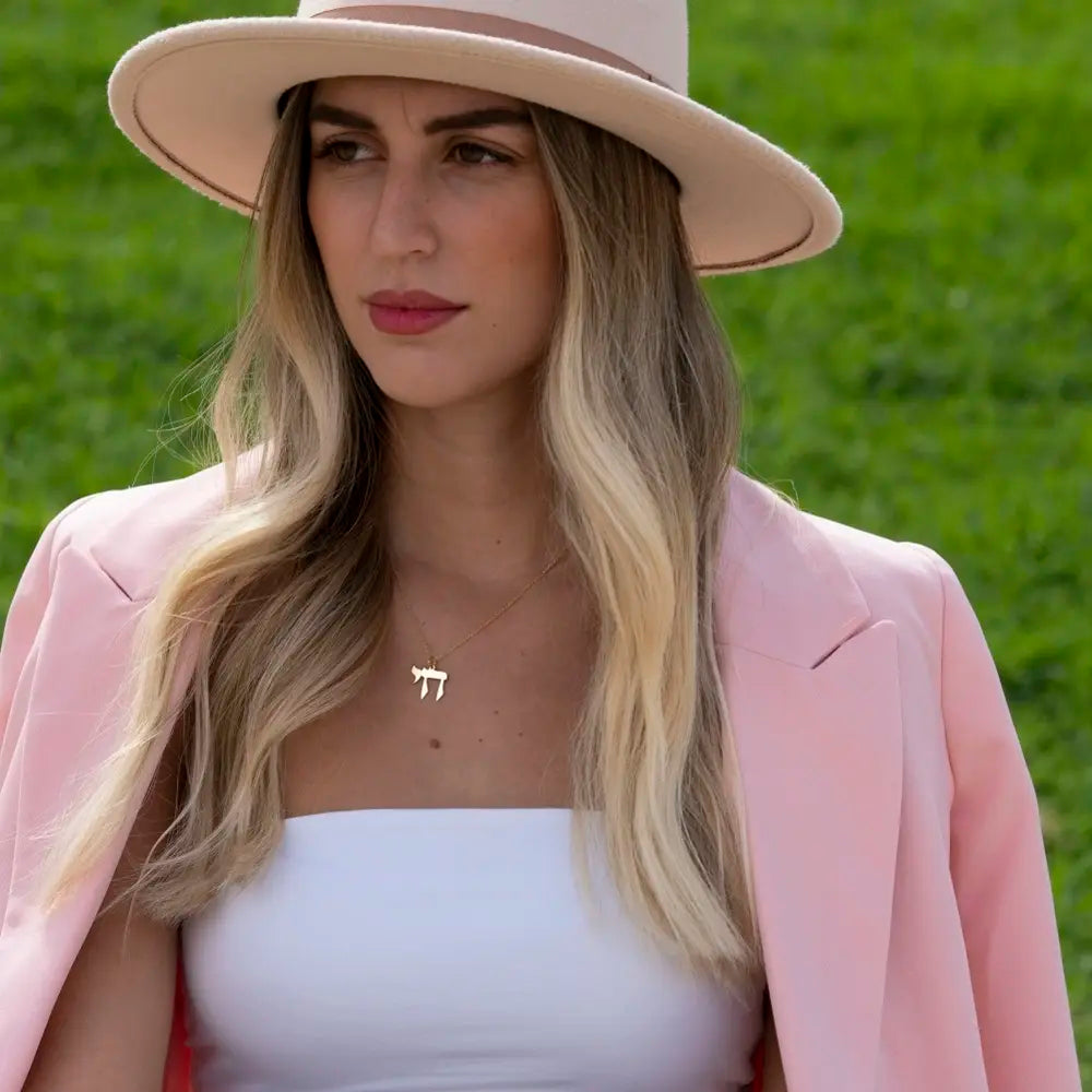 A woman wearing a light pink wide-brim hat, a matching pink blazer, and a white tube top, with a gold Hebrew letter necklace. She is photographed outdoors against a soft-focus green grass background.
