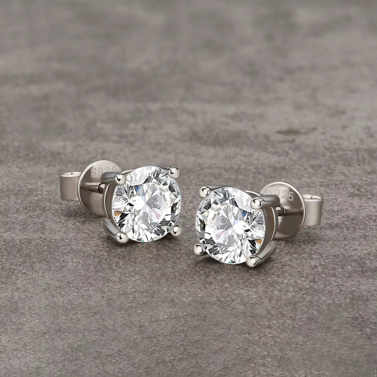 A pair of round-cut diamond stud earrings in a silver four-prong setting, displayed on a textured gray surface.
