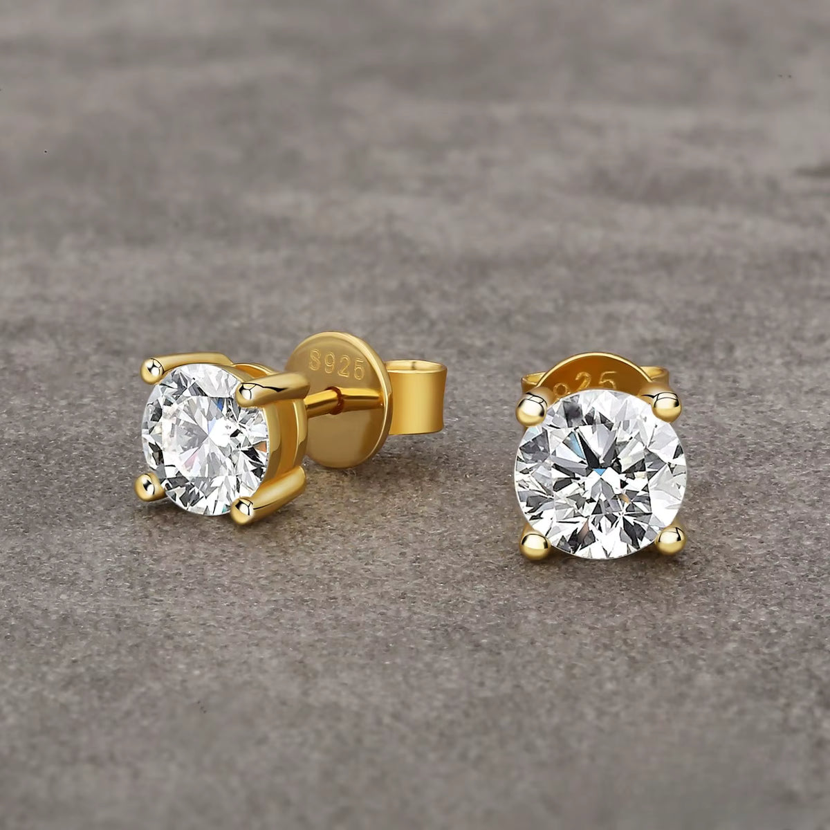 A pair of yellow gold stud earrings with round brilliant-cut clear gemstones in a four-prong setting, displayed background.