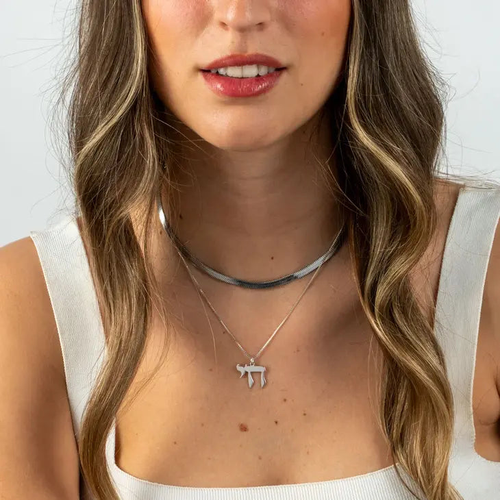 A close-up of a woman wearing a layered silver necklace set, featuring a thin choker and a delicate necklace with a Hebrew letter 'Shin' pendant, styled with a white ribbed tank top.
