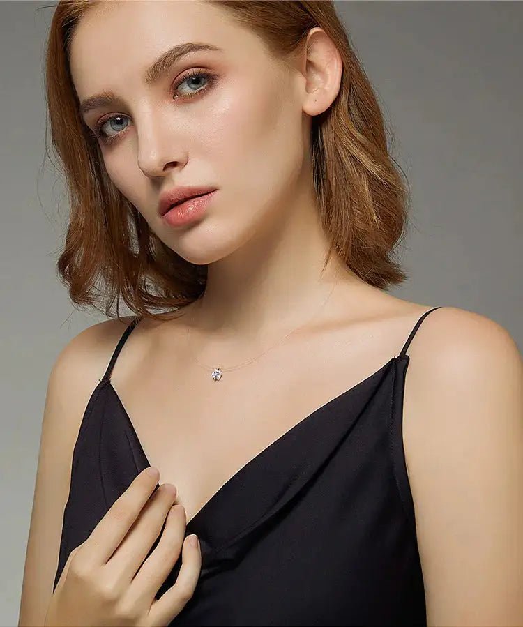 A woman with light brown hair wearing a black camisole top and a delicate silver necklace with a small clear gemstone pendant, photographed against a neutral gray background.