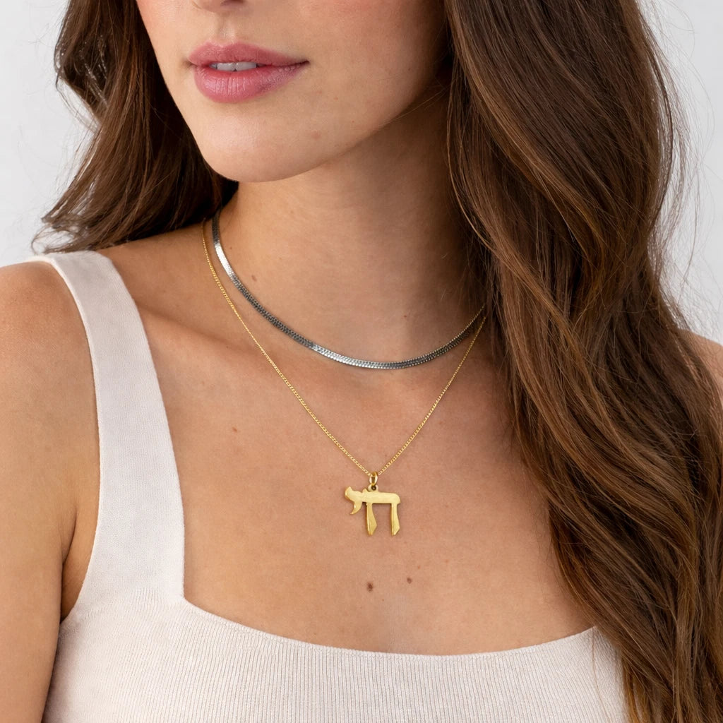 A close-up of a woman wearing layered necklaces, featuring a gold chain with a Hebrew letter Pi symbol pendant and a textured silver chain.